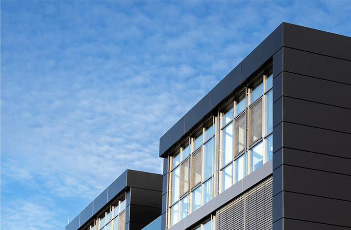 Moderne Gebäudefassade mit dunkler Verkleidung und großen Glasfenstern, aufgenommen aus der Froschperspektive vor blauem Himmel mit leichten Wolken.