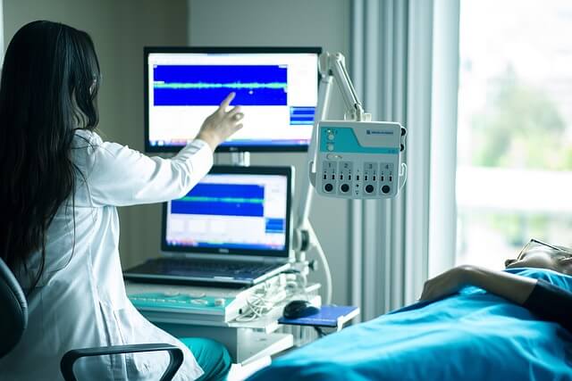 Ärztin oder medizinische Fachkraft mit langen dunklen Haaren analysiert am Monitor ein medizinisches Messdiagramm; im Vordergrund liegt eine Patientin oder ein Patient zugedeckt im Krankenhausbett.
