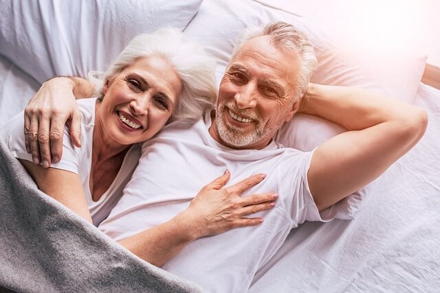 Älteres Paar mit grauen Haaren liegt lächelnd nebeneinander im Bett, beide tragen weiße T-Shirts und wirken entspannt und glücklich unter einer grauen Decke.