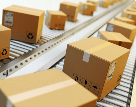 Mehrere braune Versandkartons mit Aufklebern und Warnsymbolen bewegen sich auf einem automatisierten Förderband in einer Logistikanlage.